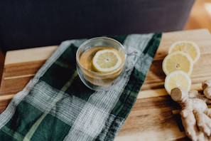 A clear glass cup filled with tea and a lemon slice on a wooden surface. Beside the cup, there is a dark green plaid cloth. To the right, there are three slices of lemon arranged next to a piece of fresh ginger.