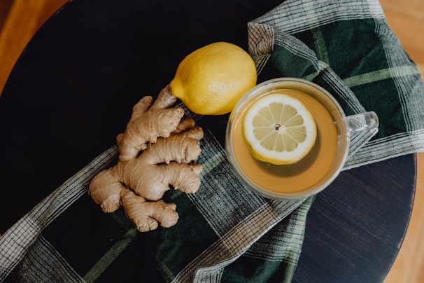 A flat lay of a ginger root and a lemon resting on a plaid cloth, next to a cup of tea with a lemon slice on its surface.