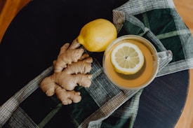 A flat lay of a ginger root and a lemon resting on a plaid cloth, next to a cup of tea with a lemon slice on its surface.