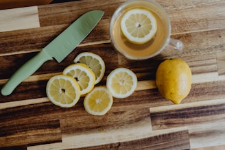 A wooden cutting board features a whole lemon, several sliced lemon pieces, a green-handled knife, and a glass mug with a lemon slice floating in a light-colored liquid.