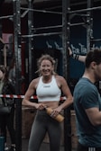 A woman in athletic wear is standing in a gym environment, smiling and holding a water bottle. She appears to be in front of gym equipment with other people around. The atmosphere looks energetic and focused.