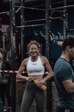 A woman in athletic wear is standing in a gym environment, smiling and holding a water bottle. She appears to be in front of gym equipment with other people around. The atmosphere looks energetic and focused.
