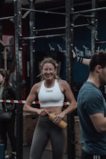 A woman in athletic wear is standing in a gym environment, smiling and holding a water bottle. She appears to be in front of gym equipment with other people around. The atmosphere looks energetic and focused.