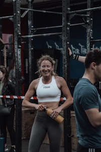 A woman in athletic wear is standing in a gym environment, smiling and holding a water bottle. She appears to be in front of gym equipment with other people around. The atmosphere looks energetic and focused.