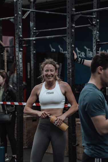 A woman in athletic wear is standing in a gym environment, smiling and holding a water bottle. She appears to be in front of gym equipment with other people around. The atmosphere looks energetic and focused.