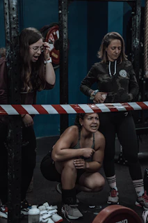 Three women are present in a gym setting. One woman is crouching and appears focused or determined, while the other two are standing nearby. The scene includes gym equipment such as a barbell plate and striped tape.