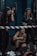 Three women are present in a gym setting. One woman is crouching and appears focused or determined, while the other two are standing nearby. The scene includes gym equipment such as a barbell plate and striped tape.