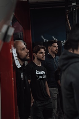 Several men stand in a gym environment, appearing focused and attentive. One of the men is wearing a black t-shirt with the word 'CrossFit' on it. The atmosphere suggests a training or group workout setting.