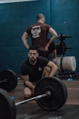 A man with glasses is crouching near a barbell on the floor, while another person with their hands on hips stands in the background, wearing a t-shirt with 'CrossFit Louvre' written on it. There are weight plates and exercise equipment visible, set against a dark teal wall.