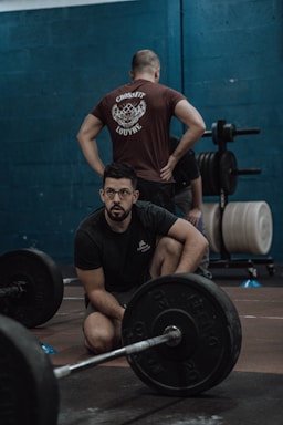 A man with glasses is crouching near a barbell on the floor, while another person with their hands on hips stands in the background, wearing a t-shirt with 'CrossFit Louvre' written on it. There are weight plates and exercise equipment visible, set against a dark teal wall.