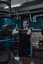 A person is lifting a heavy barbell in a gym environment. The individual is wearing a black t-shirt, black shorts with blue stripes, and wrist wraps. The background features gym equipment, a blue wall, and a board with numerous notes attached. The lighting is industrial, and various pipes and structures are visible on the ceiling.