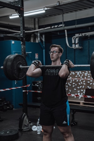 A person is lifting a heavy barbell in a gym environment. The individual is wearing a black t-shirt, black shorts with blue stripes, and wrist wraps. The background features gym equipment, a blue wall, and a board with numerous notes attached. The lighting is industrial, and various pipes and structures are visible on the ceiling.