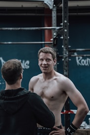 A shirtless man with short hair stands smiling next to a man facing away, both in a gym setting with workout equipment and graffiti-like writing on the wall in the background.