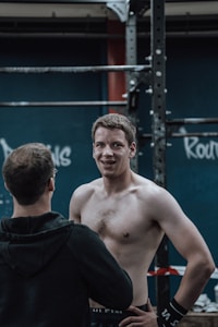 A shirtless man with short hair stands smiling next to a man facing away, both in a gym setting with workout equipment and graffiti-like writing on the wall in the background.