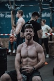 A muscular man is sitting on a bench, shirtless and smiling, in what appears to be a gym or fitness area. Other individuals are engaged in various activities in the background, including exercising and conversing. The environment suggests a communal workout space with fitness equipment and a rope in view.