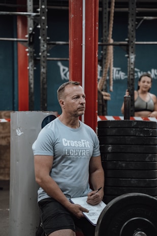 A man in a light blue CrossFit-branded shirt is sitting on a bench in a gym environment, holding a pen and a clipboard. Red gym equipment and weights are visible, with another person in workout attire blurred in the background.