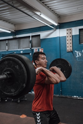 A person is performing a clean and jerk lift with a barbell in a gym setting. The individual is wearing a red shirt and black shorts. The background features blue walls and metal accents, indicating an indoor fitness environment. There are also orange cones on the floor and weightlifting equipment visible.
