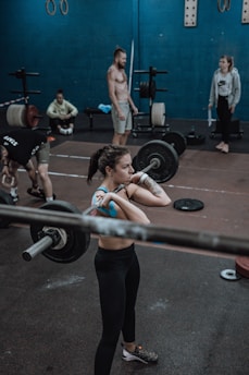 Several individuals in a gym setting are engaging in weightlifting exercises. The focus is on a woman lifting a barbell, surrounded by others who are either exercising or observing. The background shows gym equipment like weights and exercise rings.