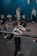 Several individuals in a gym setting are engaging in weightlifting exercises. The focus is on a woman lifting a barbell, surrounded by others who are either exercising or observing. The background shows gym equipment like weights and exercise rings.