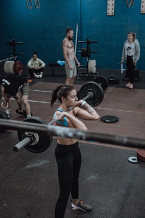 Several individuals in a gym setting are engaging in weightlifting exercises. The focus is on a woman lifting a barbell, surrounded by others who are either exercising or observing. The background shows gym equipment like weights and exercise rings.