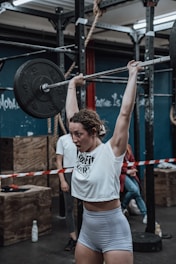 A woman is performing a weightlifting exercise in a gym. She is lifting a barbell overhead, showcasing strength and determination. The gym has a rugged, industrial feel with metal racks, ropes, and wooden boxes visible in the background. Other gym-goers are present, and a bottle of water sits on the floor.