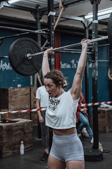 A woman is performing a weightlifting exercise in a gym. She is lifting a barbell overhead, showcasing strength and determination. The gym has a rugged, industrial feel with metal racks, ropes, and wooden boxes visible in the background. Other gym-goers are present, and a bottle of water sits on the floor.