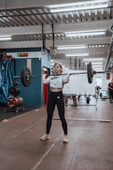 A person is performing a weightlifting exercise in a gym setting. The individual is holding a barbell with weights, standing on a brown mat. In the background, there are other gym equipment and a blue and red wall. A whiteboard with writing is visible, along with other people in the gym.