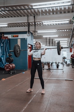 A person is performing a weightlifting exercise in a gym setting. The individual is holding a barbell with weights, standing on a brown mat. In the background, there are other gym equipment and a blue and red wall. A whiteboard with writing is visible, along with other people in the gym.