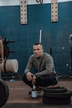 A person squats in a gym setting, with a focused expression. The background includes gym equipment such as weights and various gear. The person is holding a bottle of sparkling water and wearing a gray long-sleeve shirt and athletic shoes. The wall is painted dark teal, and gym equipment like weight plates are visible.