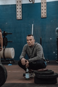 A person squats in a gym setting, with a focused expression. The background includes gym equipment such as weights and various gear. The person is holding a bottle of sparkling water and wearing a gray long-sleeve shirt and athletic shoes. The wall is painted dark teal, and gym equipment like weight plates are visible.