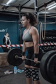 A woman in athletic clothing is performing a deadlift with a barbell in a gym setting. She is wearing a grey sports bra, black shorts, knee sleeves with a swirl pattern, and wrist straps. The gym has industrial style equipment, including ropes and weight racks in the background. Another person is seen in the background, standing near a barricade.