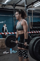A woman in athletic clothing is performing a deadlift with a barbell in a gym setting. She is wearing a grey sports bra, black shorts, knee sleeves with a swirl pattern, and wrist straps. The gym has industrial style equipment, including ropes and weight racks in the background. Another person is seen in the background, standing near a barricade.