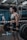 A woman in athletic clothing is performing a deadlift with a barbell in a gym setting. She is wearing a grey sports bra, black shorts, knee sleeves with a swirl pattern, and wrist straps. The gym has industrial style equipment, including ropes and weight racks in the background. Another person is seen in the background, standing near a barricade.