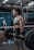 A woman in athletic clothing is performing a deadlift with a barbell in a gym setting. She is wearing a grey sports bra, black shorts, knee sleeves with a swirl pattern, and wrist straps. The gym has industrial style equipment, including ropes and weight racks in the background. Another person is seen in the background, standing near a barricade.