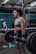A woman in athletic clothing is performing a deadlift with a barbell in a gym setting. She is wearing a grey sports bra, black shorts, knee sleeves with a swirl pattern, and wrist straps. The gym has industrial style equipment, including ropes and weight racks in the background. Another person is seen in the background, standing near a barricade.