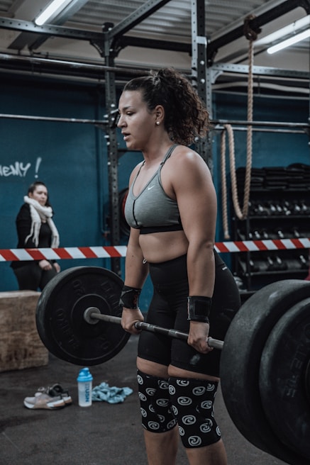 A woman in athletic clothing is performing a deadlift with a barbell in a gym setting. She is wearing a grey sports bra, black shorts, knee sleeves with a swirl pattern, and wrist straps. The gym has industrial style equipment, including ropes and weight racks in the background. Another person is seen in the background, standing near a barricade.