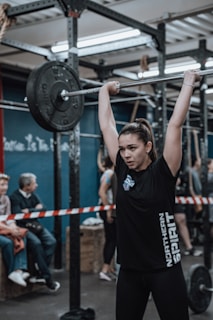 A young woman is lifting a barbell with heavy weights above her head in a gym setting. She appears focused and determined. Several people are present in the background, some sitting and observing. The space is an industrial-style gym with metal racks and equipment. The atmosphere is competitive and energetic.