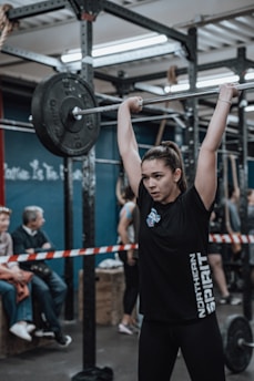 A young woman is lifting a barbell with heavy weights above her head in a gym setting. She appears focused and determined. Several people are present in the background, some sitting and observing. The space is an industrial-style gym with metal racks and equipment. The atmosphere is competitive and energetic.