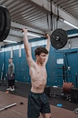 A person is performing an overhead weightlifting exercise in a gym. The setting has gym equipment such as barbells, weights, and rubber flooring. Another individual stands in the background observing or ready to assist.