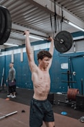 A person is performing an overhead weightlifting exercise in a gym. The setting has gym equipment such as barbells, weights, and rubber flooring. Another individual stands in the background observing or ready to assist.