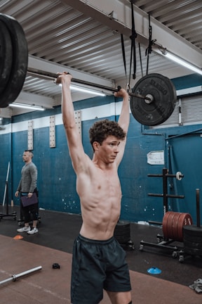 A person is performing an overhead weightlifting exercise in a gym. The setting has gym equipment such as barbells, weights, and rubber flooring. Another individual stands in the background observing or ready to assist.