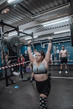 A person is engaged in a weightlifting activity in a gym setting, lifting a barbell overhead. Several individuals in the background are observing, with gym equipment and a rugged industrial ceiling visible.