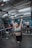 A person is engaged in a weightlifting activity in a gym setting, lifting a barbell overhead. Several individuals in the background are observing, with gym equipment and a rugged industrial ceiling visible.