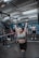 A person is engaged in a weightlifting activity in a gym setting, lifting a barbell overhead. Several individuals in the background are observing, with gym equipment and a rugged industrial ceiling visible.