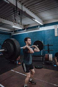 A person is in a gym performing a barbell clean exercise. The gym has blue walls and is equipped with various workout equipment, including weight plates and gymnastic rings hanging from the ceiling. The individual is wearing a dark t-shirt and shorts, focusing on lifting the barbell.