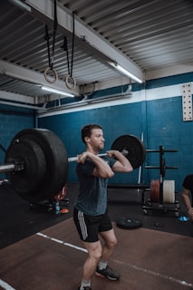 A person is in a gym performing a barbell clean exercise. The gym has blue walls and is equipped with various workout equipment, including weight plates and gymnastic rings hanging from the ceiling. The individual is wearing a dark t-shirt and shorts, focusing on lifting the barbell.
