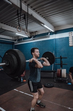 A person is in a gym performing a barbell clean exercise. The gym has blue walls and is equipped with various workout equipment, including weight plates and gymnastic rings hanging from the ceiling. The individual is wearing a dark t-shirt and shorts, focusing on lifting the barbell.