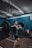 A person is in a gym performing a barbell clean exercise. The gym has blue walls and is equipped with various workout equipment, including weight plates and gymnastic rings hanging from the ceiling. The individual is wearing a dark t-shirt and shorts, focusing on lifting the barbell.