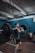 A person is in a gym performing a barbell clean exercise. The gym has blue walls and is equipped with various workout equipment, including weight plates and gymnastic rings hanging from the ceiling. The individual is wearing a dark t-shirt and shorts, focusing on lifting the barbell.