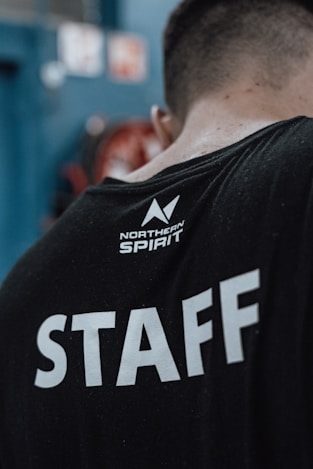 A person wearing a black t-shirt with 'STAFF' written in large white letters on the back. Above it, there is a smaller logo with the words 'Northern Spirit'. The person's head is turned away from the camera, showing only the back of their head and upper body. The background is blurred, with hints of blue and red colors.